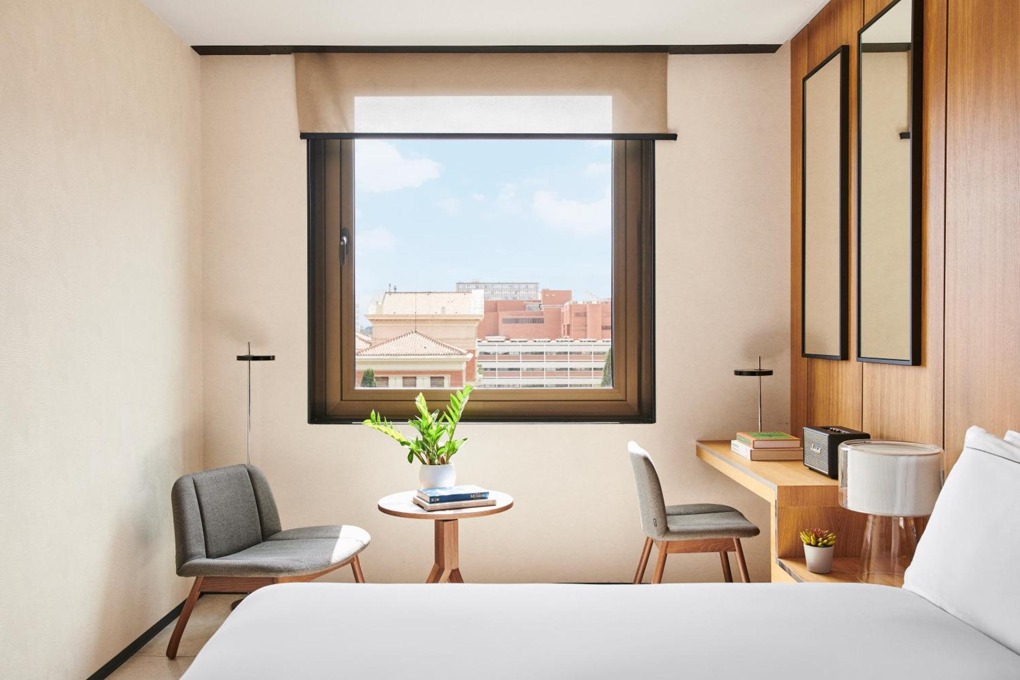 Grand Hyatt Barcelona Ξενοδοχείο Εξωτερικό φωτογραφία The photo depicts a modern hotel room with a cozy and minimalist design. There's a large window allowing natural light to enter, showcasing a view of the outside cityscape. In the room, two chairs are positioned near a small round table, which has a 
